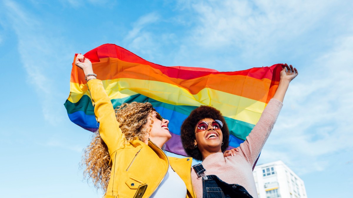 Zwei Freund*innen, die eine Pride-Flagge hochhalten und glücklich nach vorne blicken.