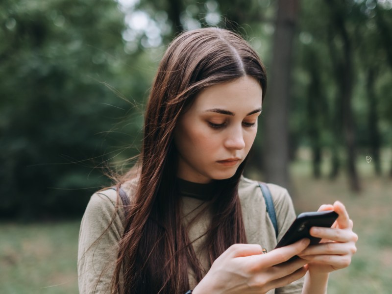 Frau auf einer Wiese mit Bäumen im Hintergrund, die nachdenklich auf ihr Handy runterschaut.
