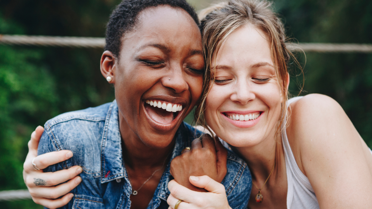 Zwei Frauen auf einer Bank draußen, die sich umarmen und lachen.