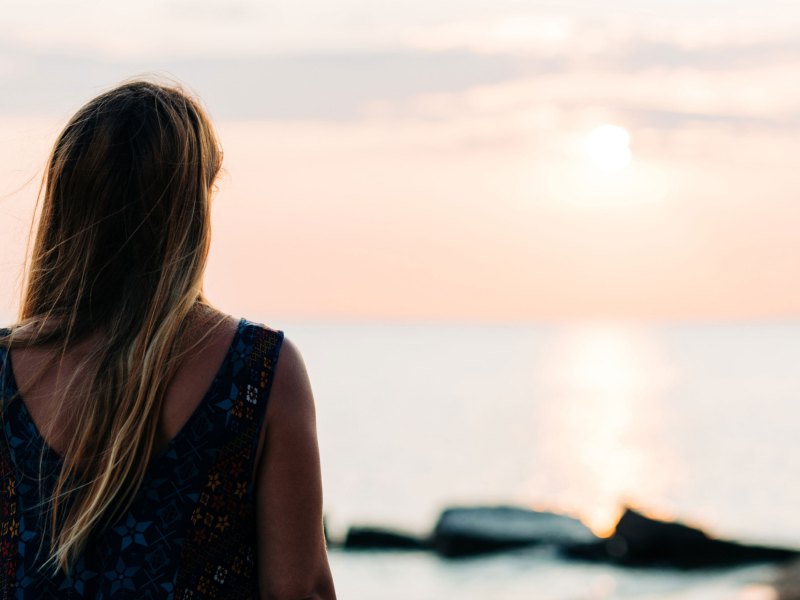 Frau steht am Meer und schaut den Sonnenuntergang