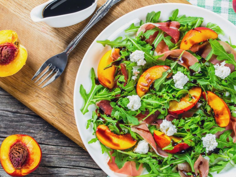 Ruculasalat mit Pfirsichen und Burrata auf einem großen weißen Teller.