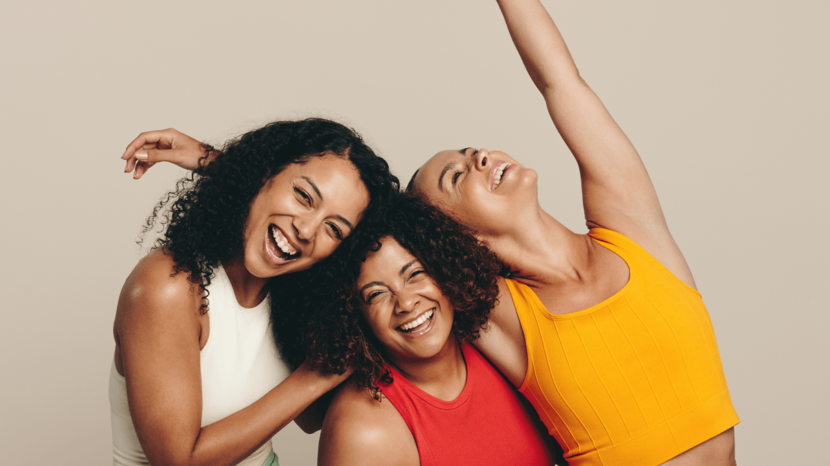 Drei Frauen vor einer hellbraunen Wand, die Sportklamotten anhaben und gemeinsam lachen.