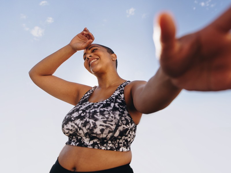 Junge plus-Size Frau in gemustertem Sport-BH lacht und streckt bei sonnigem Wetter die Hand in die Kamera vor einem klaren Himmel.