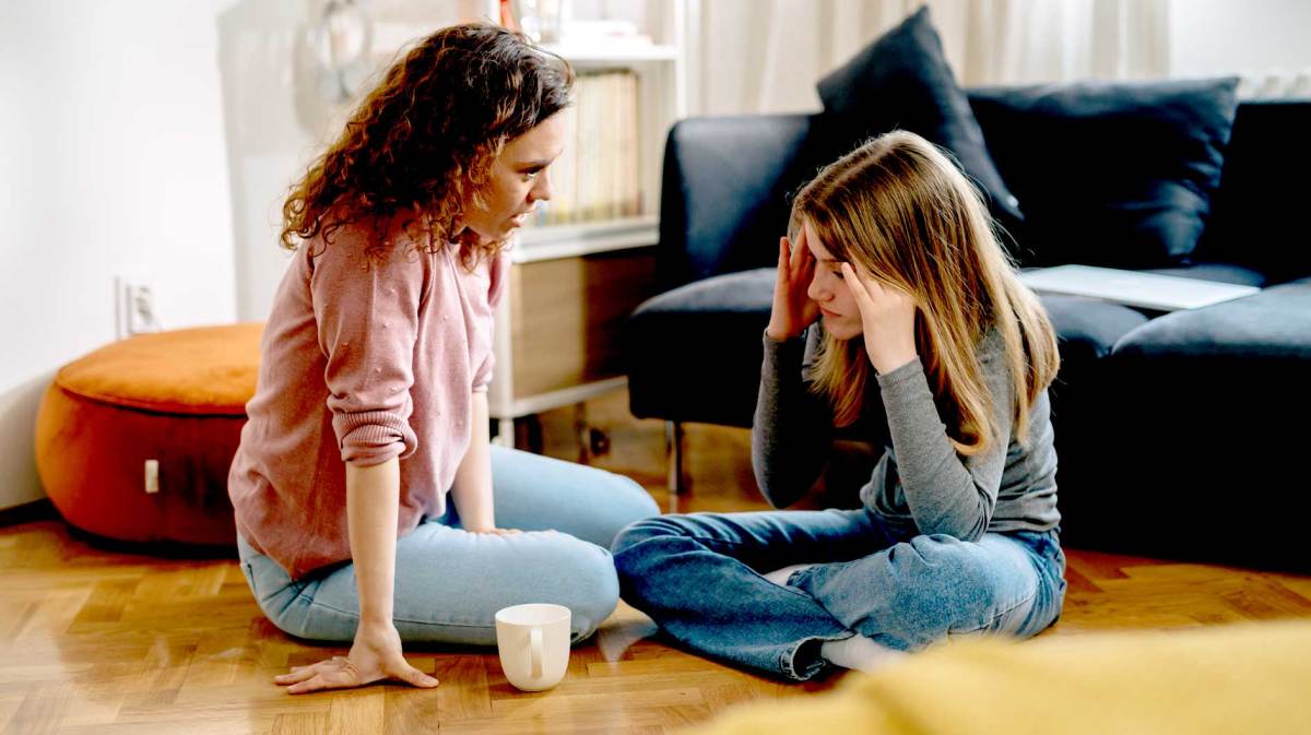 Mutter und Tochter sitzen im Wohnzimmer auf dem Fußboden und unterhalten sich. Die Tochter sieht genervt aus.
