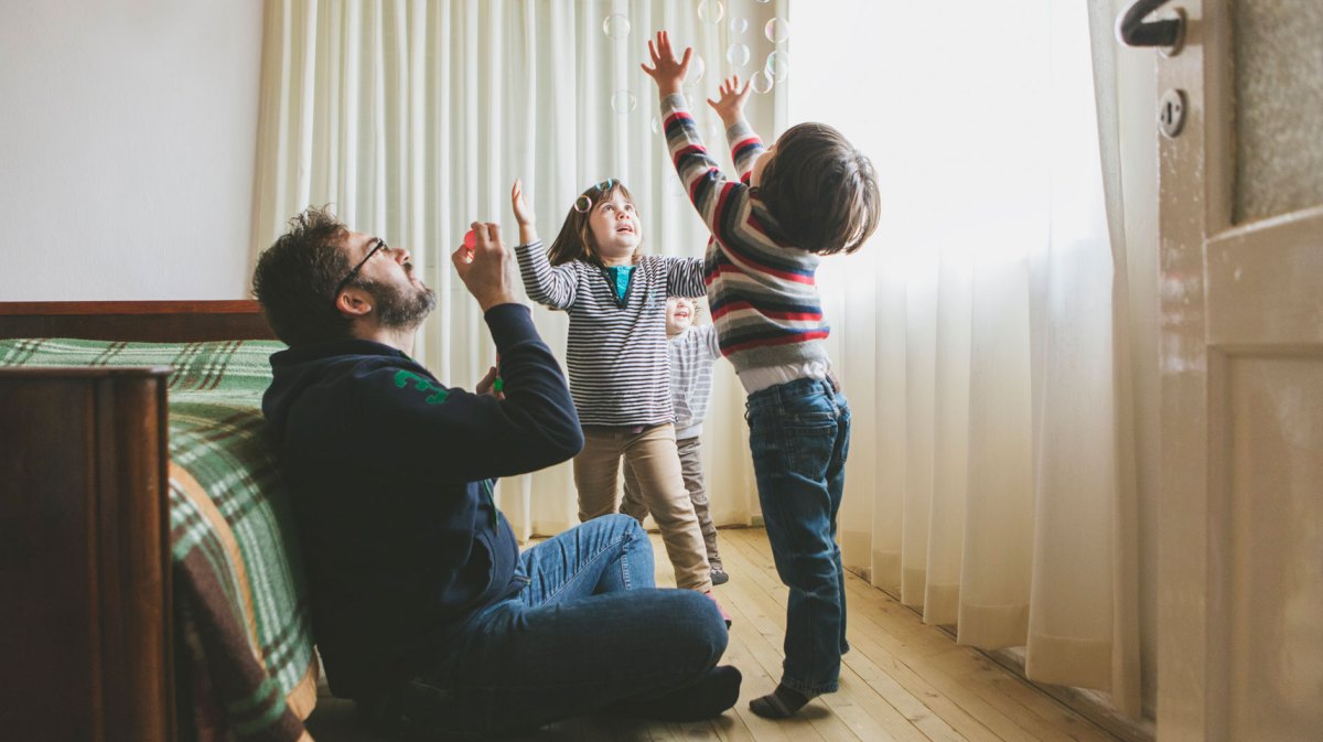 Vater sitzt auf dem Boden im Schlafzimmer und pustet Seifenblasen. Seine drei kleinen Kinder versuchen, diese zu fangen.
