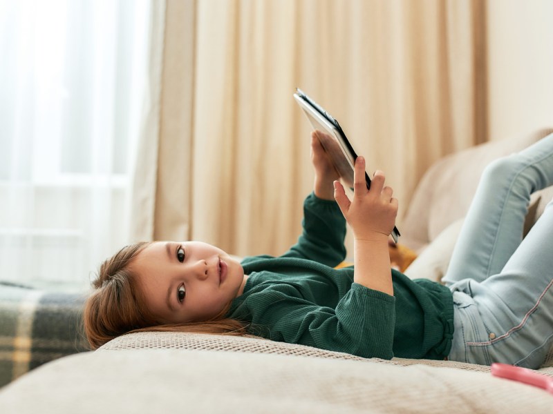 Mädchen, ca. 6 Jahre alt, liegt mit Tablet auf dem Sofa, die Beine in die Luft gestreckt.
