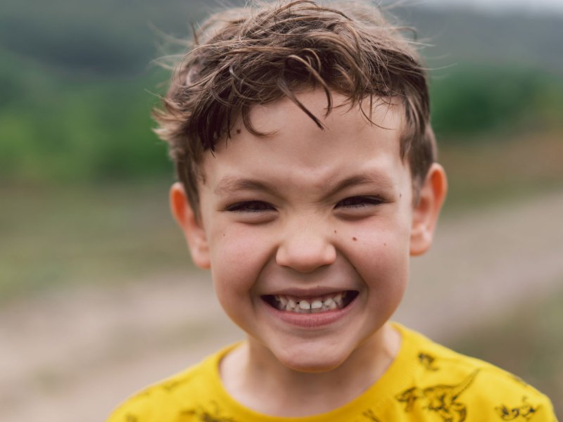 Porträt eines Jungen draußen, ca. 6 Jahre als, der in die Kamera grinst.