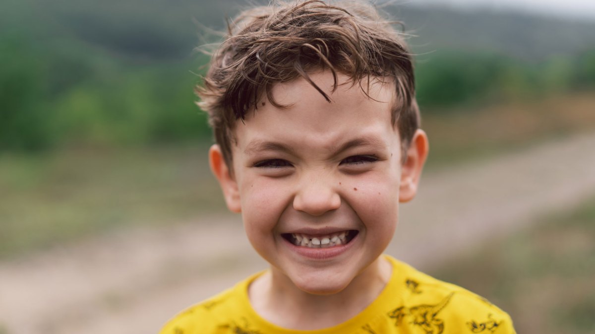 Porträt eines Jungen draußen, ca. 6 Jahre als, der in die Kamera grinst.