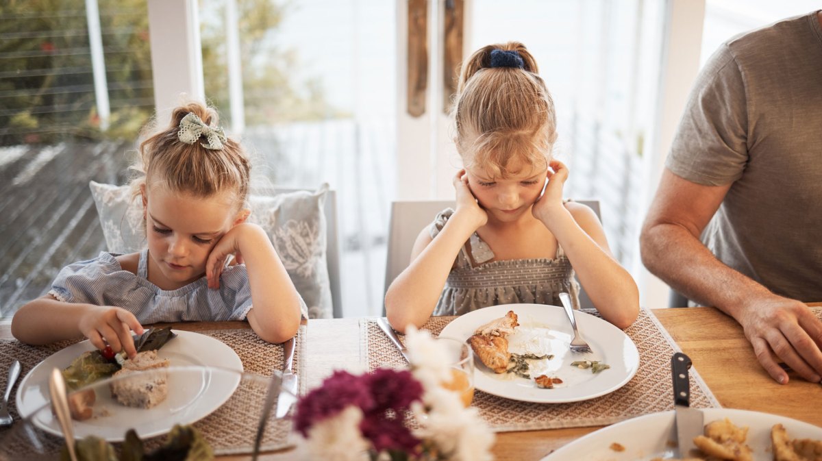 Zwei Mädchen sitzen am Familientisch und schauen das Essen auf ihren Tellern skeptisch an.