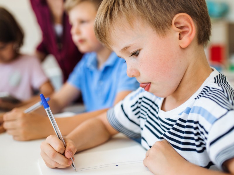 Ein Grundschüler malt etwas hoch konzentriert auf ein Blatt Papier. Neben ihm sitzen weitere Schüler*innen und steht eine Lehrerin.
