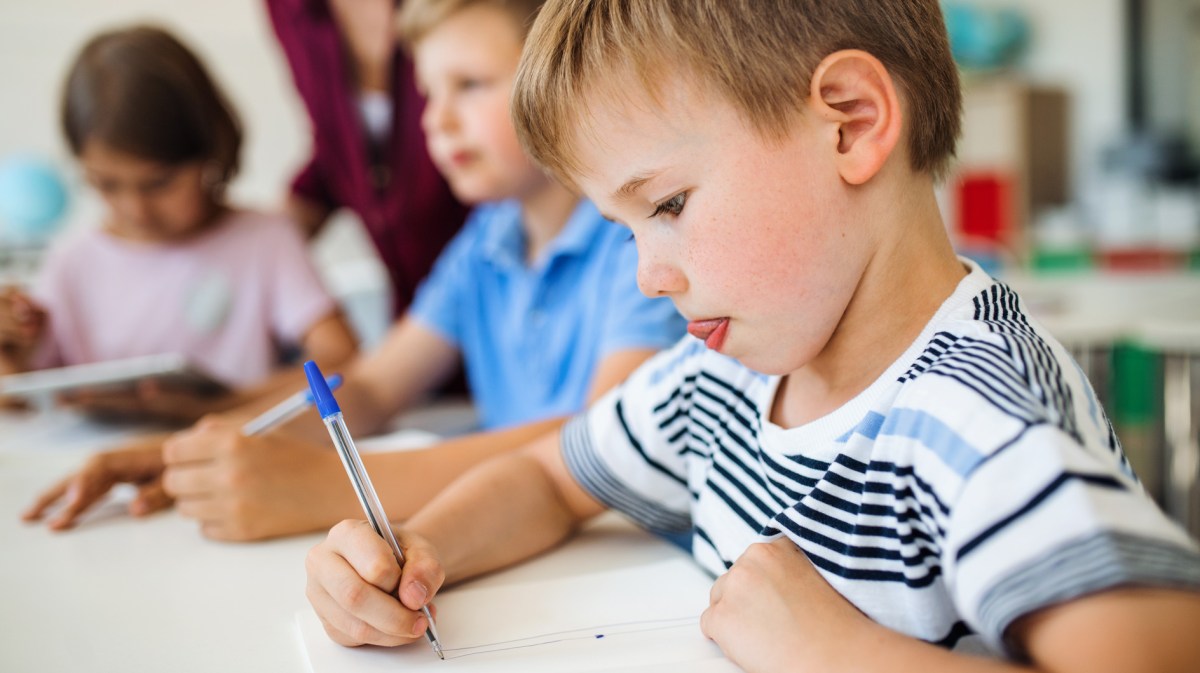 Ein Grundschüler malt etwas hoch konzentriert auf ein Blatt Papier. Neben ihm sitzen weitere Schüler*innen und steht eine Lehrerin.