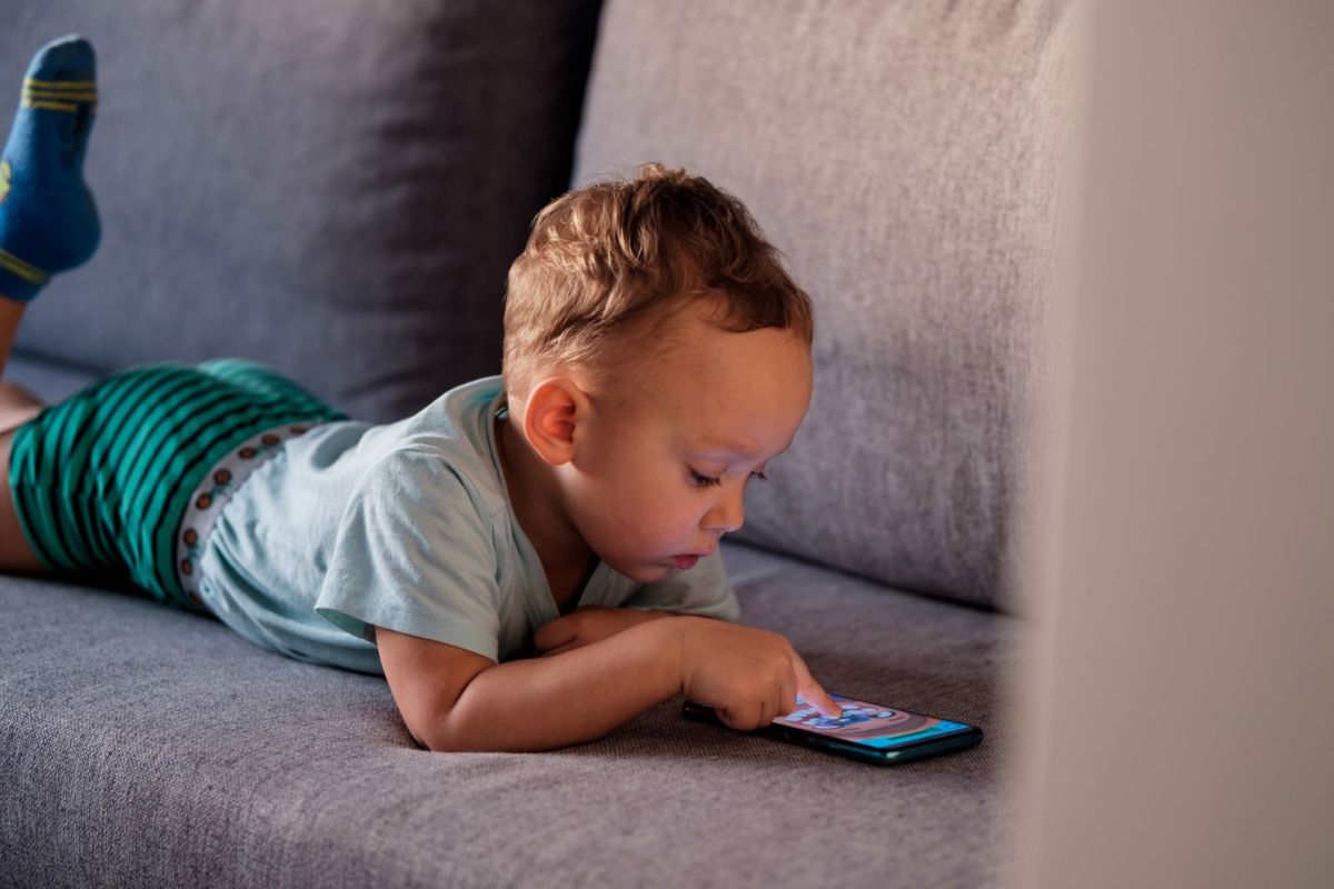 Kleinkind liegt auf dem Sofa und schaut auf ein Handy