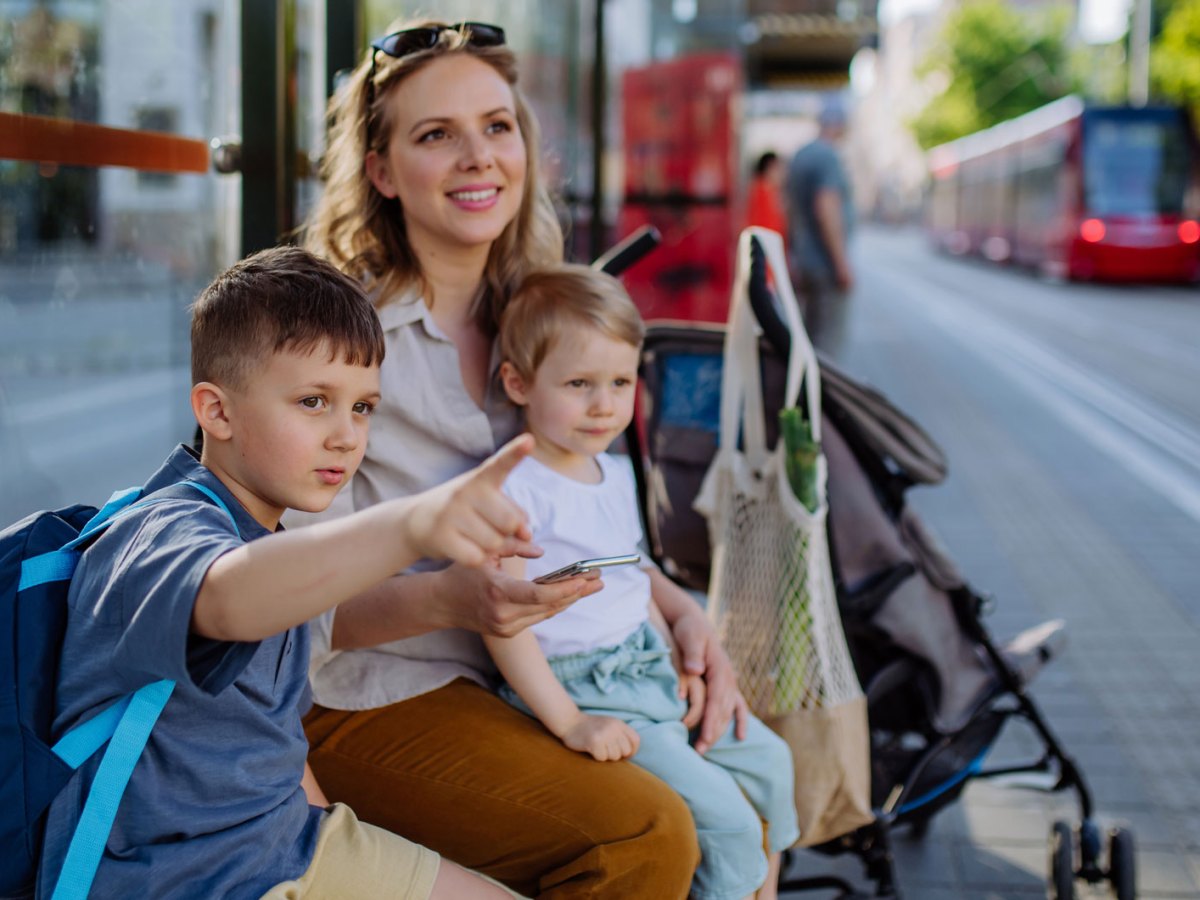Mutter und ihre zwei Kinder sitzen an einem schönen Frühlingstag an einer Bahnhaltestelle, warten geduldig und schauen sich währenddessen um.