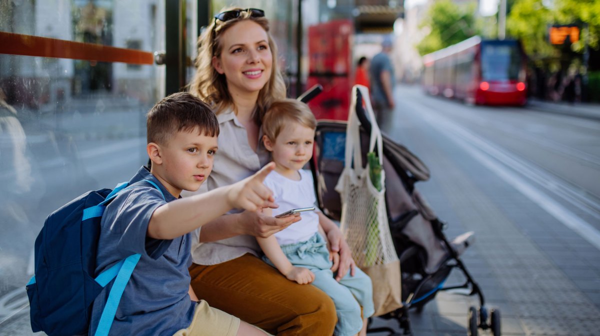 Mutter und ihre zwei Kinder sitzen an einem schönen Frühlingstag an einer Bahnhaltestelle, warten geduldig und schauen sich währenddessen um.