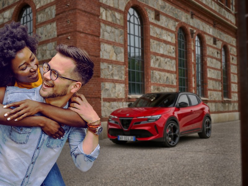 Ein lächelnder Mann mit Brille trägt eine lachende Frau auf dem Rücken, beide vor einem alten Gebäude mit rotem SUV im Hintergrund.