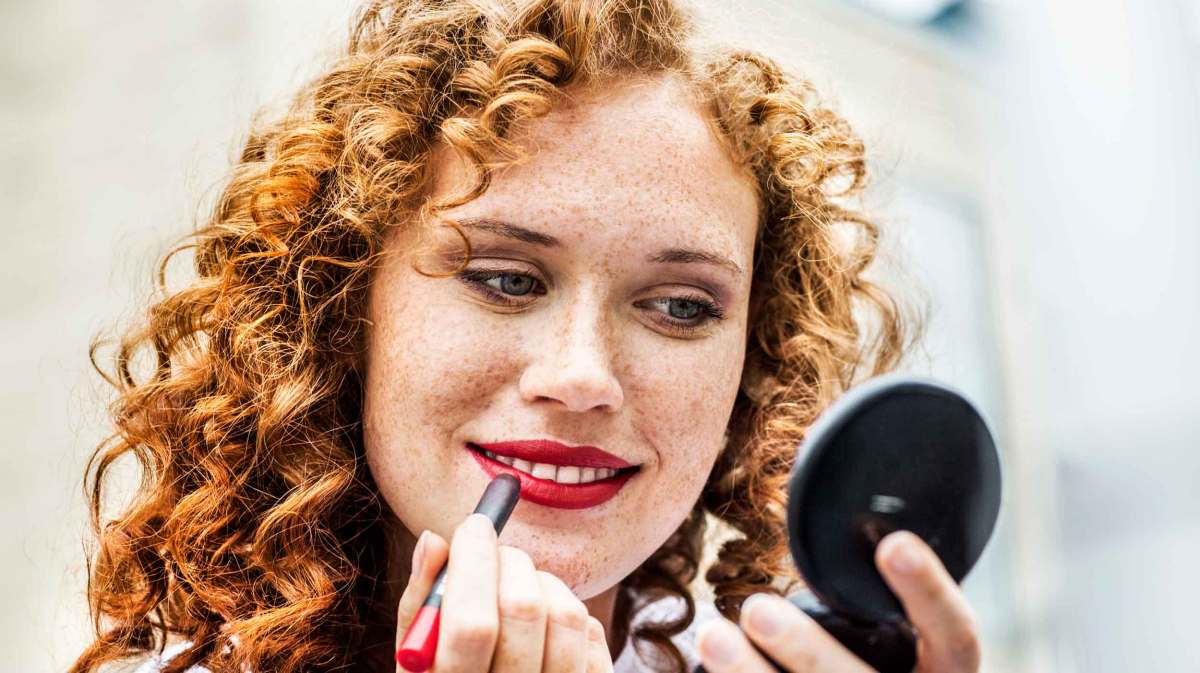 Frau mit roten Locken schminkt sich die Lippen mit einem roten Lippenstift.