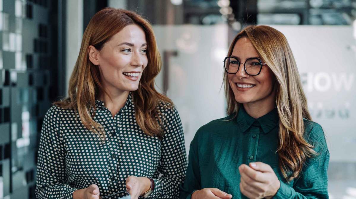 Zwei erfolgreiche junge Frauen mit Businesskleidung gehen durch das Büro und unterhalten sich angeregt.