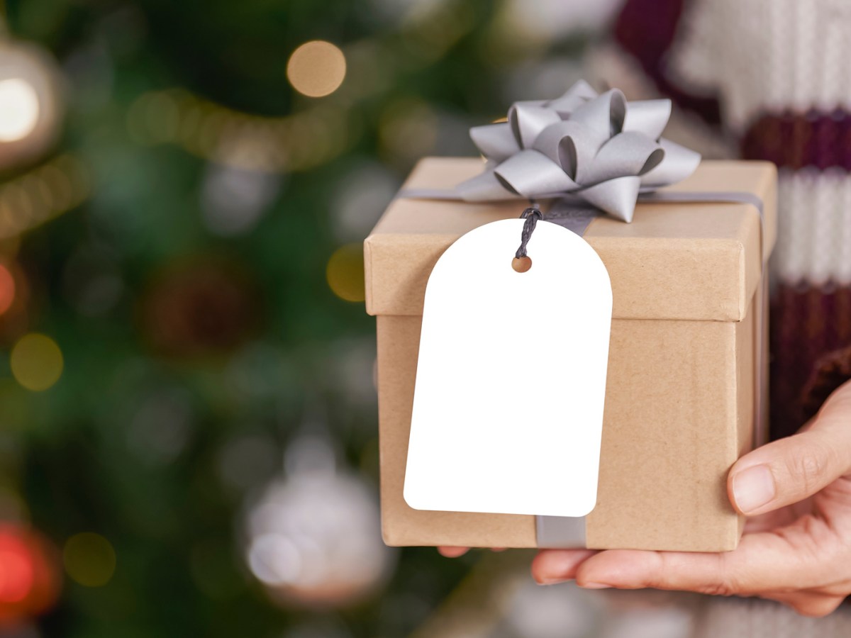 Hände halten ein Weihnachtspäckchen, im Hintergrund sieht man den Tannenbaum verschwommen.