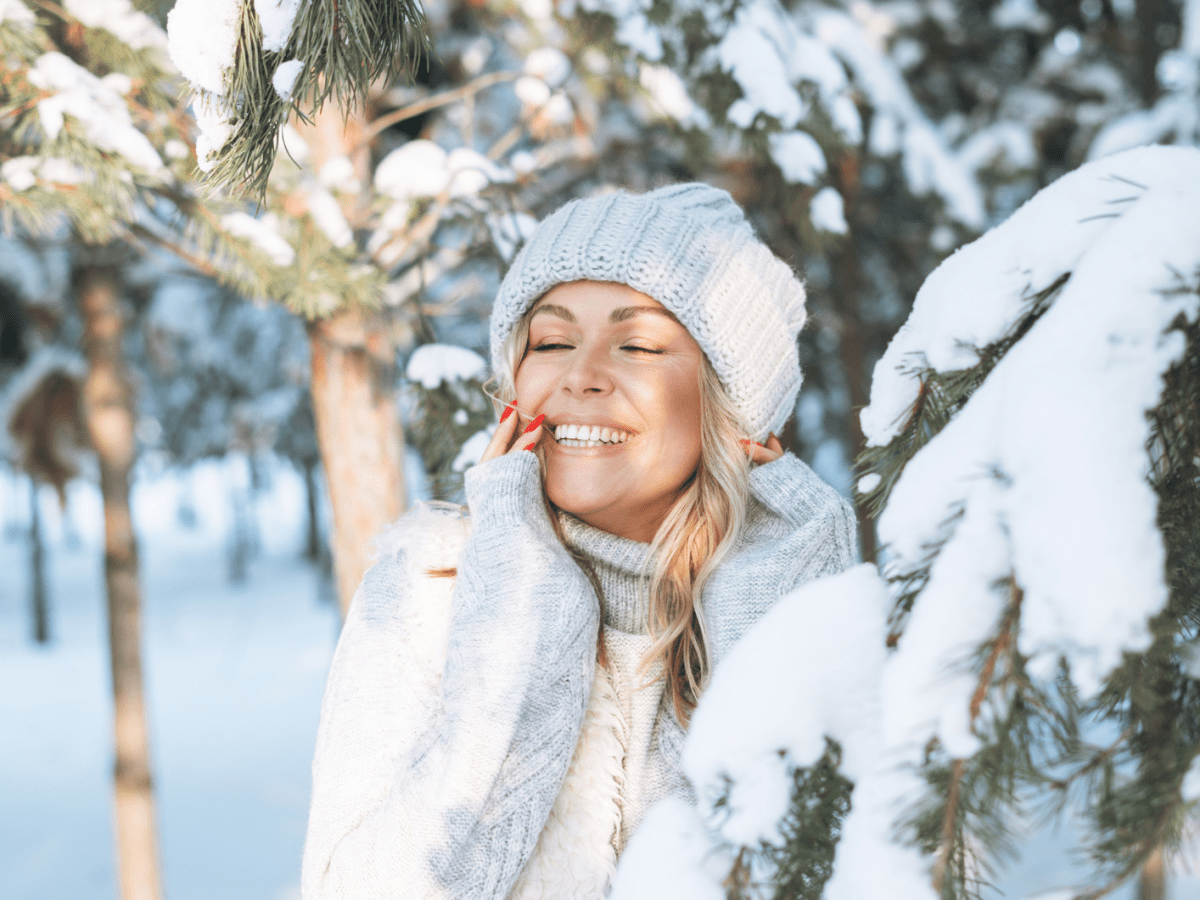 Junge Frau mit strahlendem Lächeln und rotem Nagellack lehnt sich im warmen Winter-Outfit an einen schneebedeckten Baum.