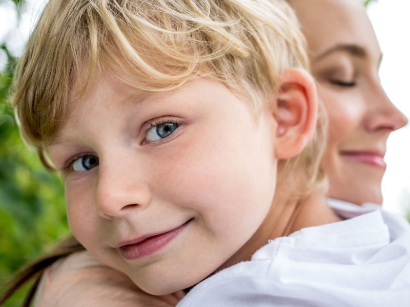 Porträt eines Jungen, ca. 6 Jahre alt, der sich glücklich an die Schulter seiner Mutter lehnt, die ihn scheinbar trägt.