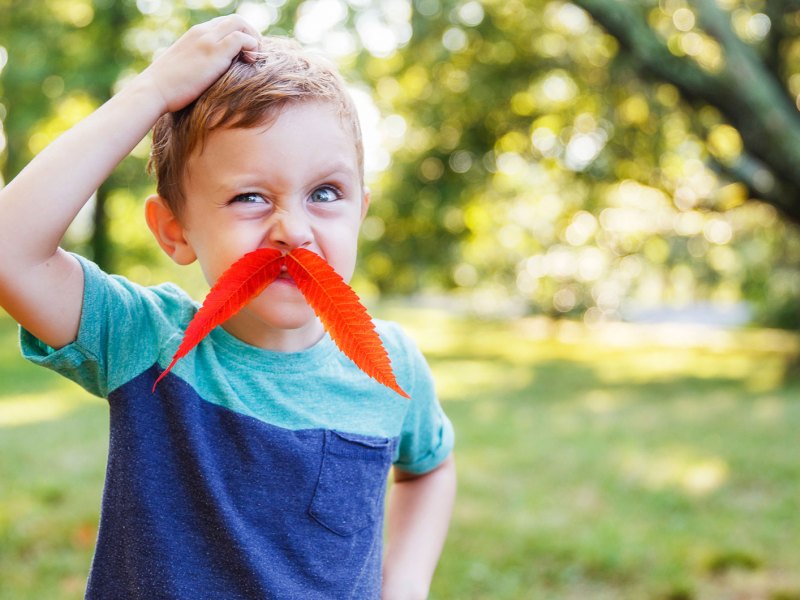 Junge, ca. 5 Jahre, albert draußen herum. Er hat sich ein Blatt als Schnurrbart aufgeklebt.