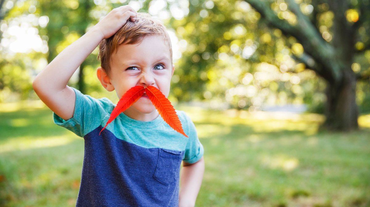 Junge, ca. 5 Jahre, albert draußen herum. Er hat sich ein Blatt als Schnurrbart aufgeklebt.