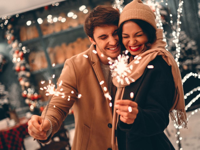 Mann und Frau in Winterbekleidung vor weihnachtlich geschmücktem Haus, mit Wunderkerzen in der Hand.