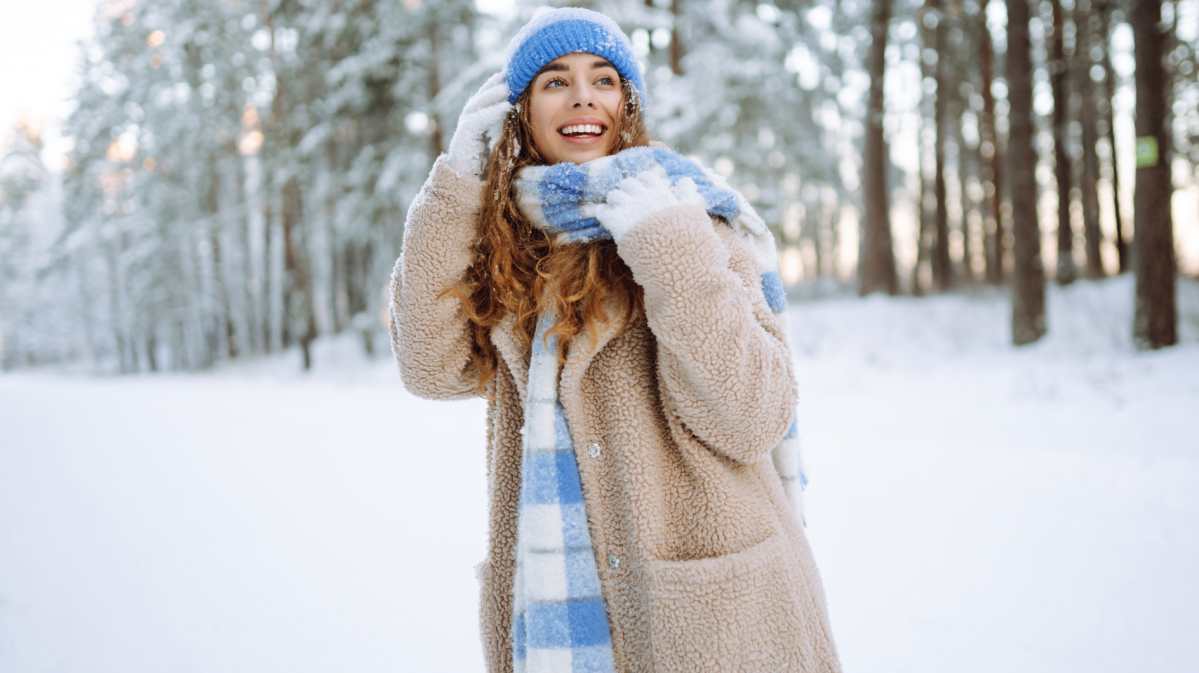 Junge Frau mit braunen Locken und blauer Mütze lacht fröhlich im verschneiten Winterwald, eingehüllt in einen beigen Mantel und karierten Schal.