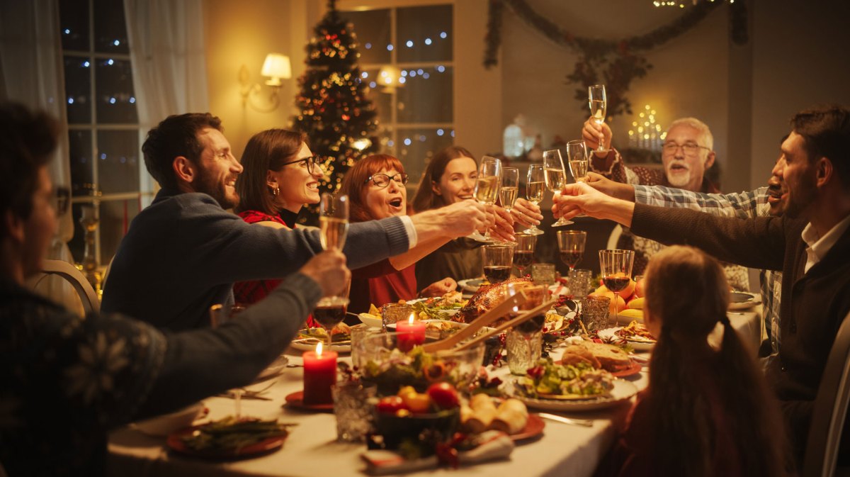 Familie und Freunde sitzen an einem festlich gedeckten Tisch, im Hintergrund leuchtet der Weihnachtsbaum und alle prosten sich zu.