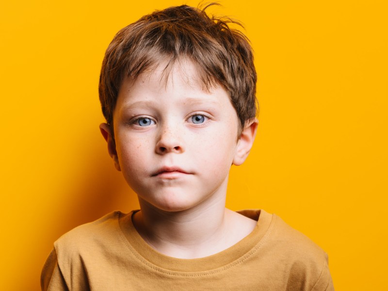 Porträt eines Jungen, ca. 7 Jahre alt, der in einem gelben Shirt vor einer gelben Wand steht und ernst schaut.