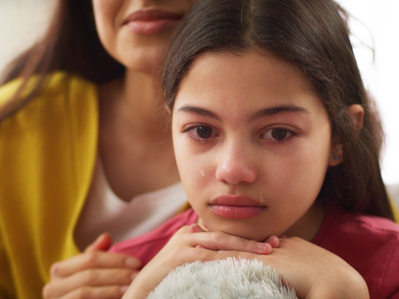 Nahaufnahme eines Mädchens, ca. 12 Jahre alt, das seinen Kopf auf dem Knie abgestützt hat und weint. Im Hintergrund ist seine Mutter verschwommen zu erkennen, die ihr eine Hand auf die Schulter gelegt hat.