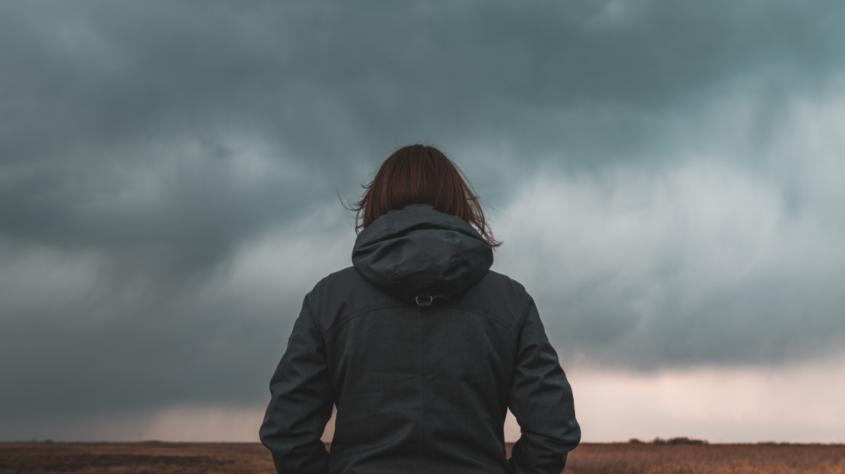 Frau steht mit dem Rücken zur Kamera vor einem düsteren Wolkenhimmel