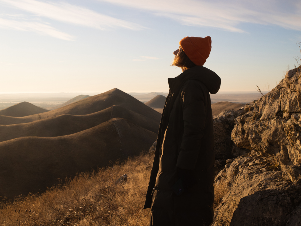 Eine Person mit orangefarbener Mütze und langem Mantel steht bei Sonnenuntergang auf einem Felsvorsprung und blickt über hügelige Landschaft.