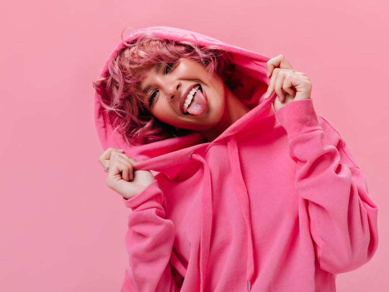Frau mit pinken Kapuzenpulli, vor einer punken Wand mit pinken Haaren, die ihre Zunge herausstreckt und lächelt.