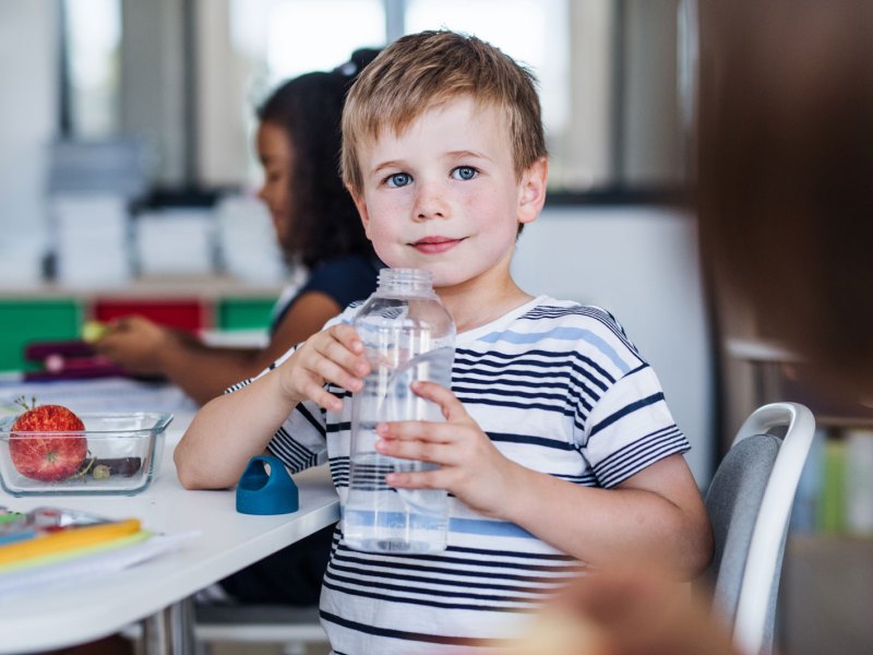 Schuljunge im Klassenraum hält eine große Wasserflasche in seinen Händen und hat sich seitlich zur Kamera gedreht.