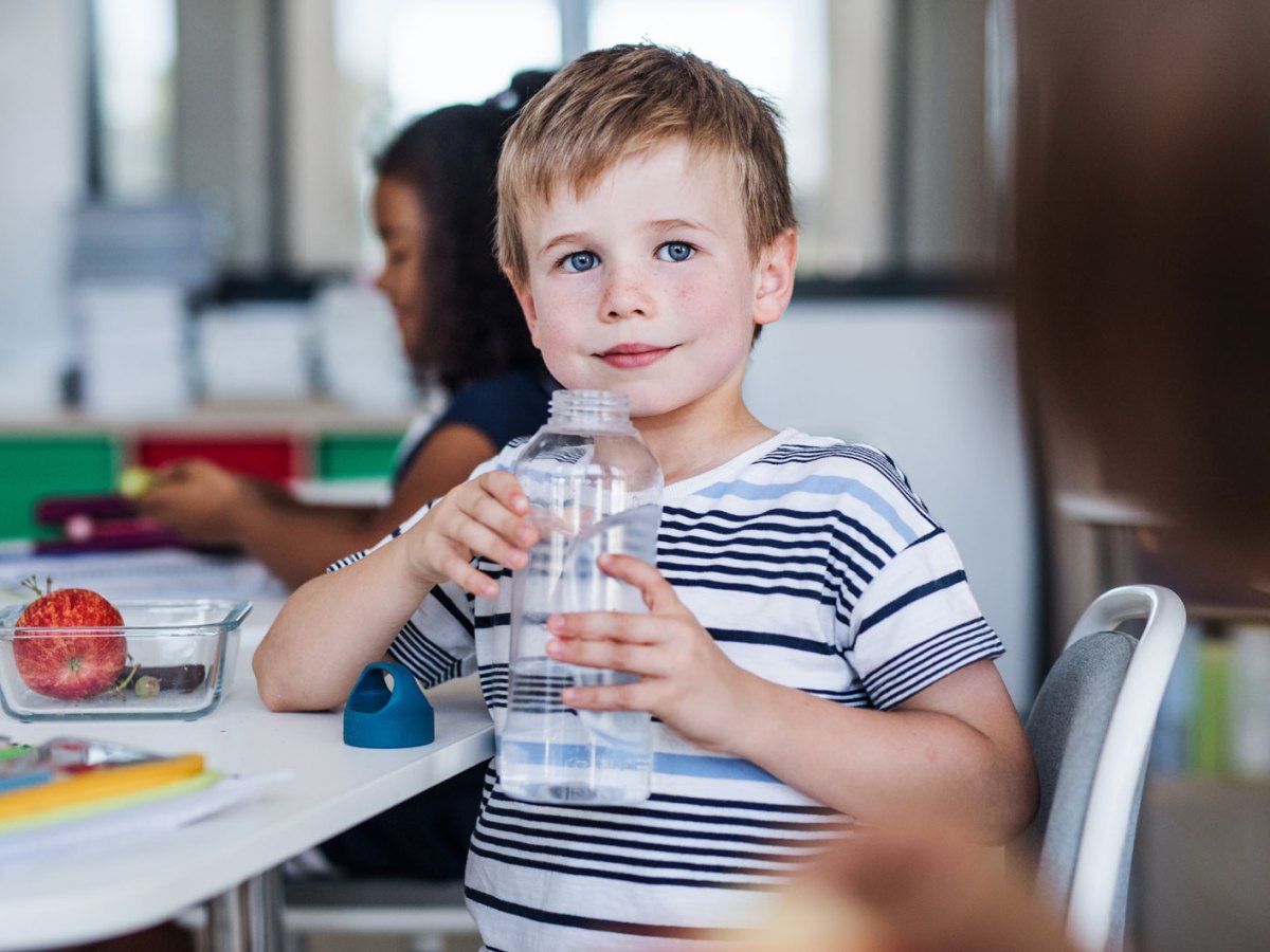 Schuljunge im Klassenraum hält eine große Wasserflasche in seinen Händen und hat sich seitlich zur Kamera gedreht.