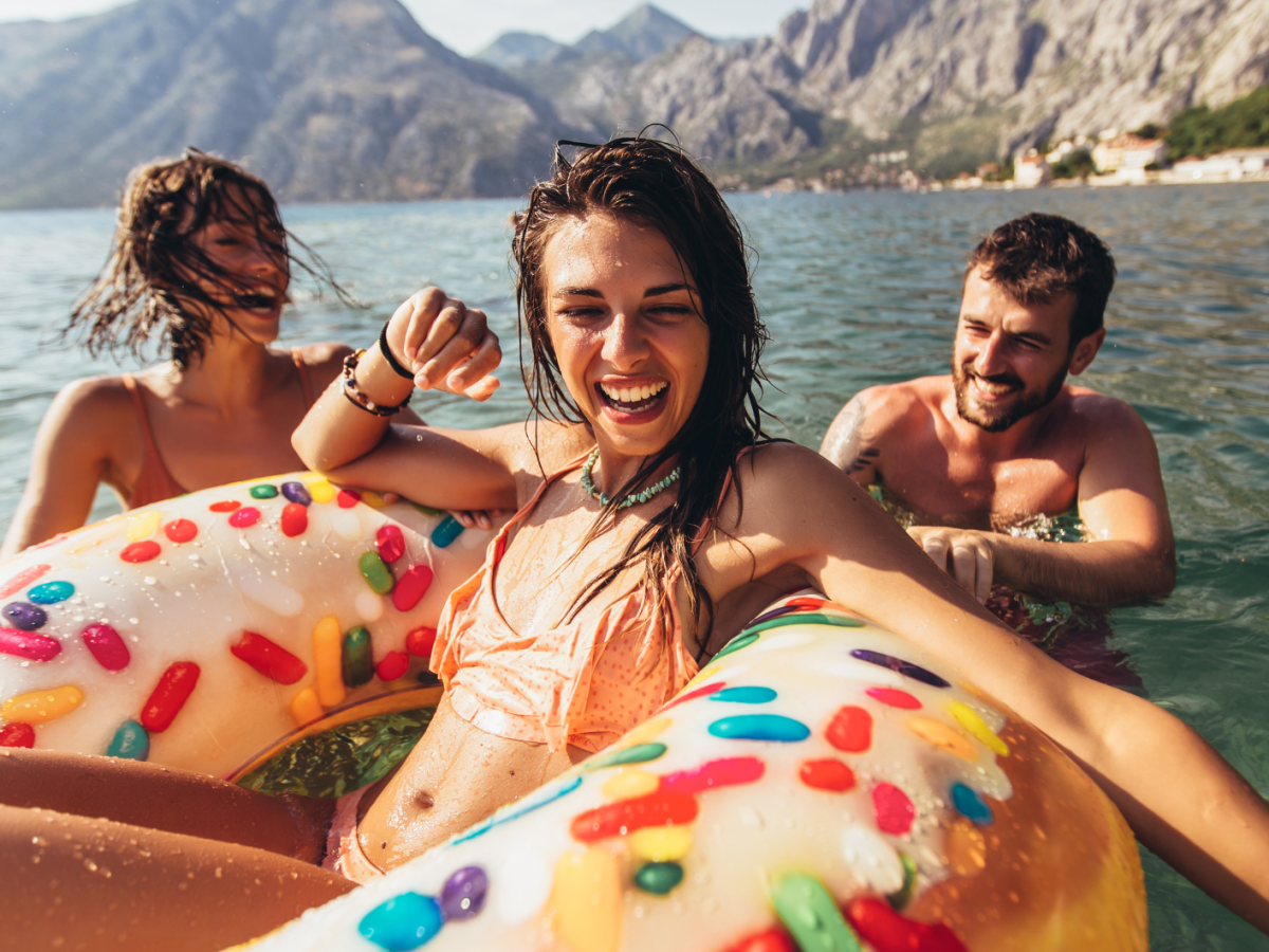 Zwei Frauen und ein Mann lachend im Wasser mit einem Schwimmreifen vor einer Bergkulisse
