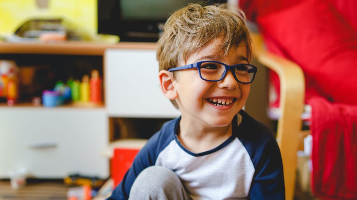 Porträt eines Jungen, ca. 7 Jahre alt, der auf dem Boden seines Kinderzimmers sitzt, umgeben von Spielsachen und glücklich lacht.