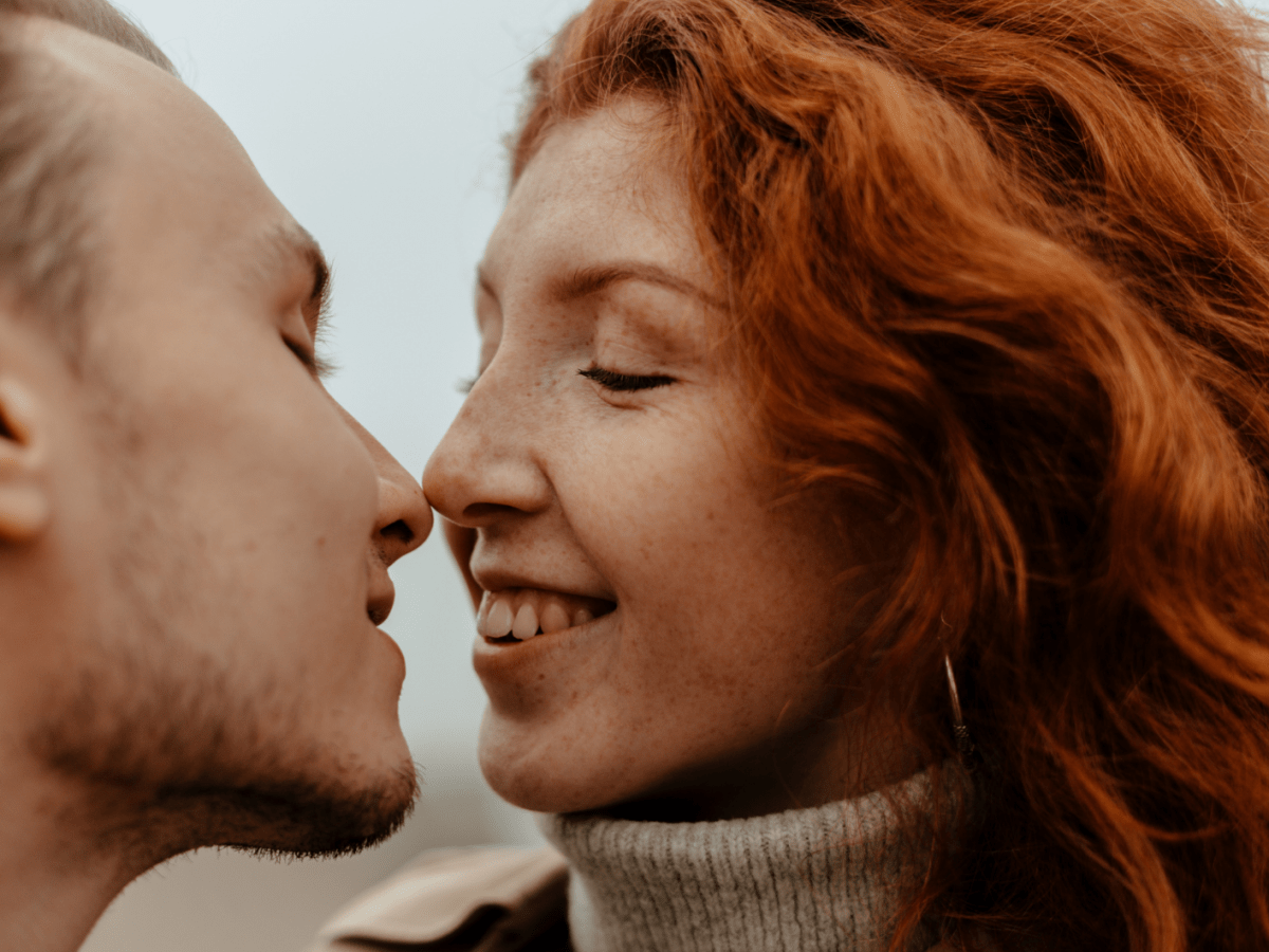 Frau mit roten Haaren küsst einen Mann mit dunklen, kurzen Haaren und lächelt.