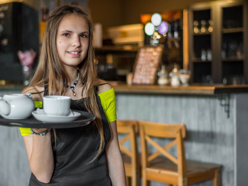 Mädchen im Teenageralter in einem Kaffee. Es trägt eine Schürze und ein Tablett.