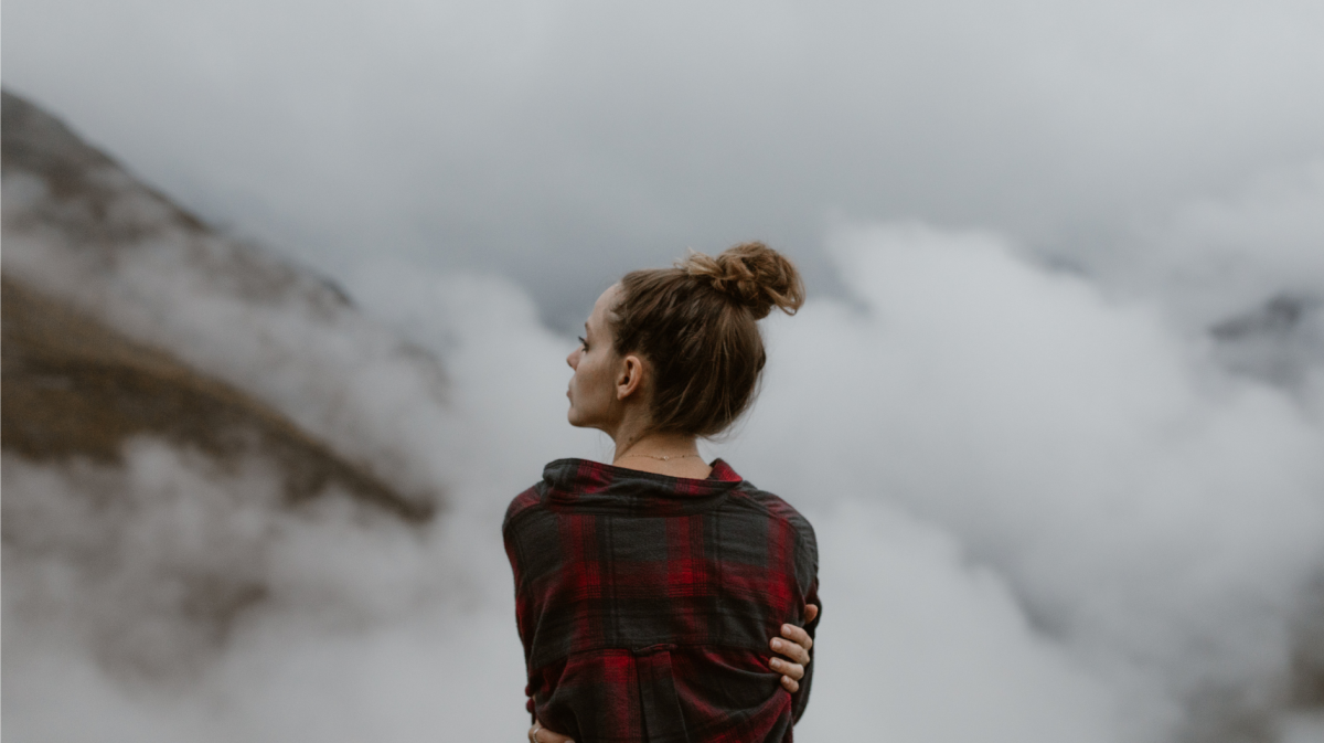 Frau mit Dutt auf einem Berg im Nebel, die die Arme um sich schlingt und nachdenklich zur Seite blickt.