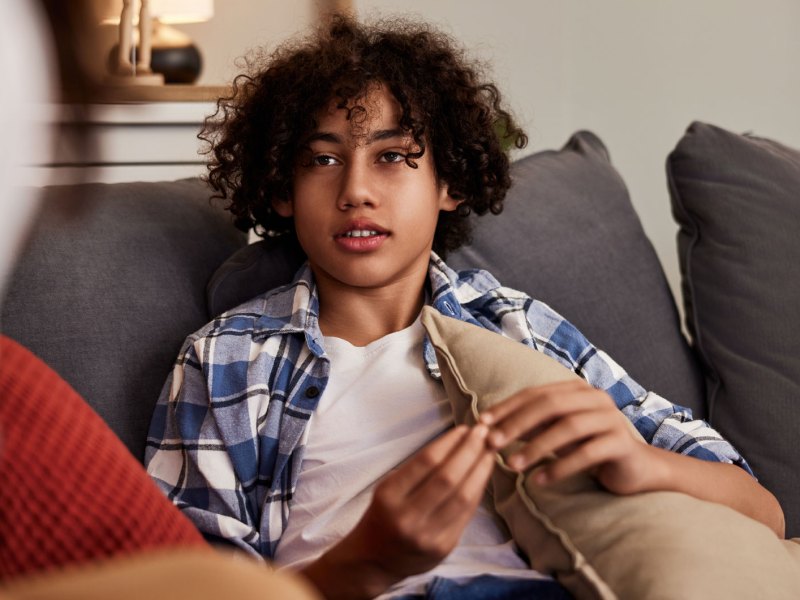 Porträt eines Teenagers, der auf einer Couch sitzt, ein Kissen festhält und ernst mit jemandem spricht.