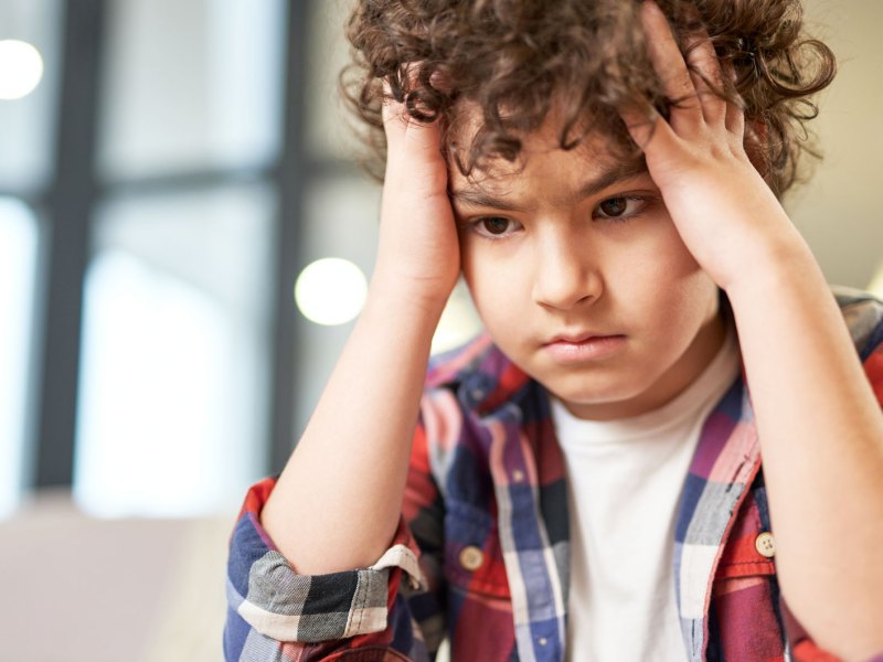 Junge, ca. 11 Jahre, sitzt am Tisch, hält sich den Kopf und schaut verzweifelt nach unten.