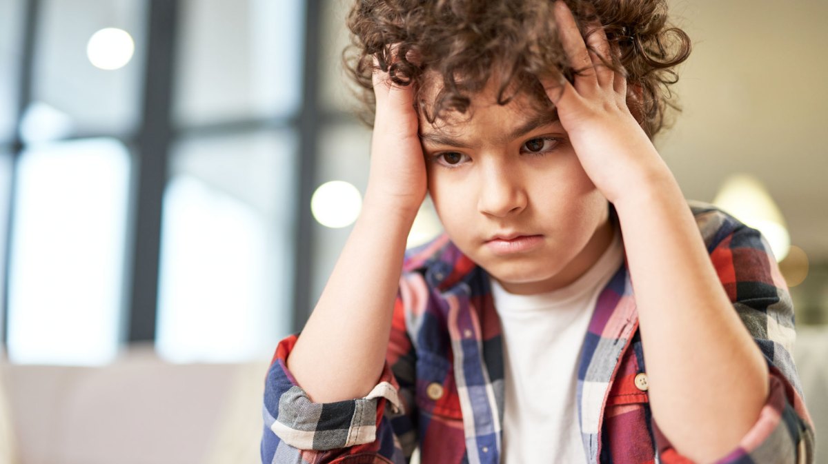 Junge, ca. 11 Jahre, sitzt am Tisch, hält sich den Kopf und schaut verzweifelt nach unten.