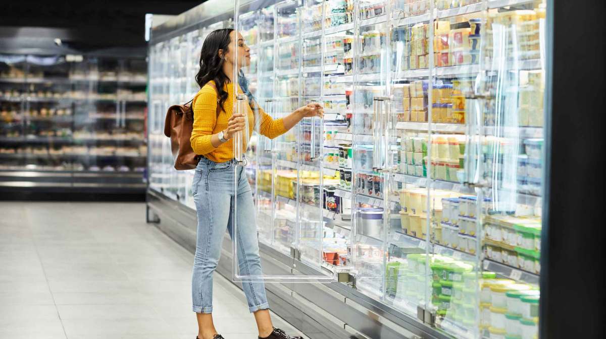 Eine junge Frau mit langen schwarzen Haaren und gelbem Pullover greift in einem Supermarkt nach einem Produkt aus einer Kühltheke.