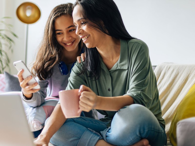 Mutter und Tochter sitzen gemütlich auf dem Sofa, und schauen beide lachend auf den Bildschirm des Laptops.