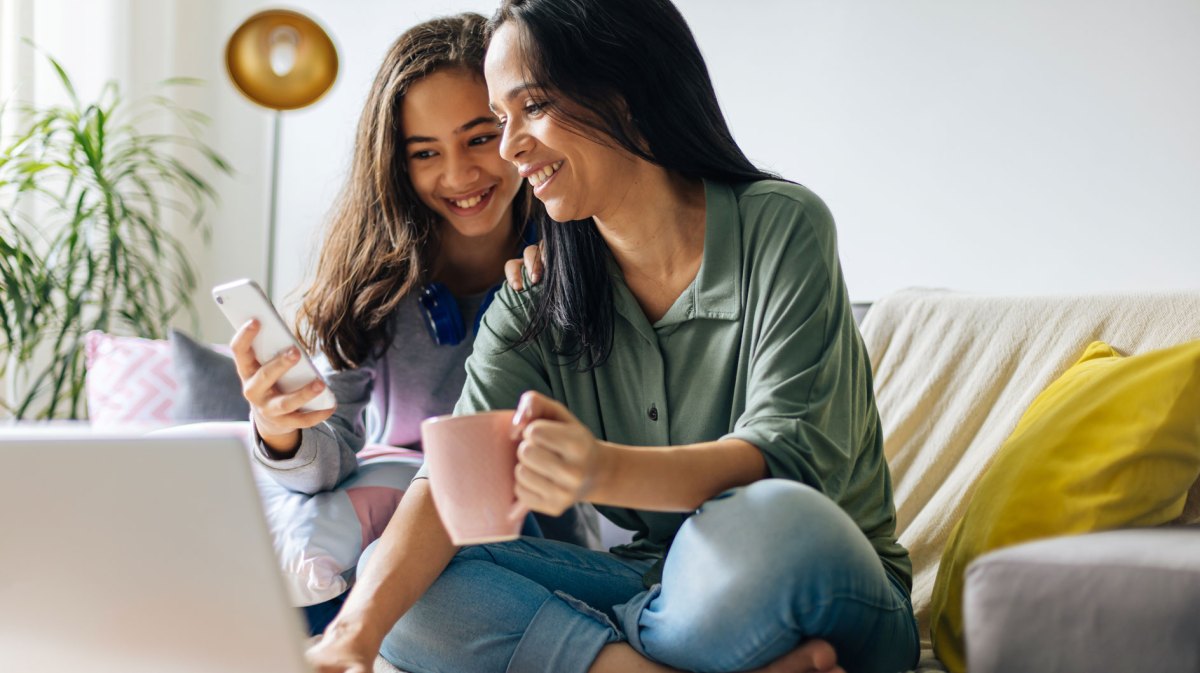 Mutter und Tochter sitzen gemütlich auf dem Sofa, und schauen beide lachend auf den Bildschirm des Laptops.