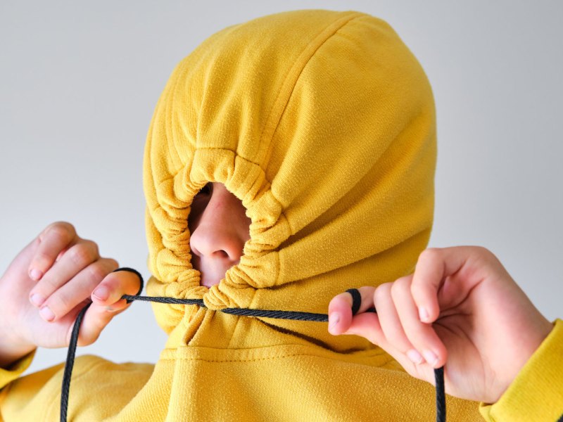 Porträt eines Jungen im Teenageralter, der die Kapuze seines gelben Pullis so weit zu zieht, dass nur noch die Nase herausschaut.
