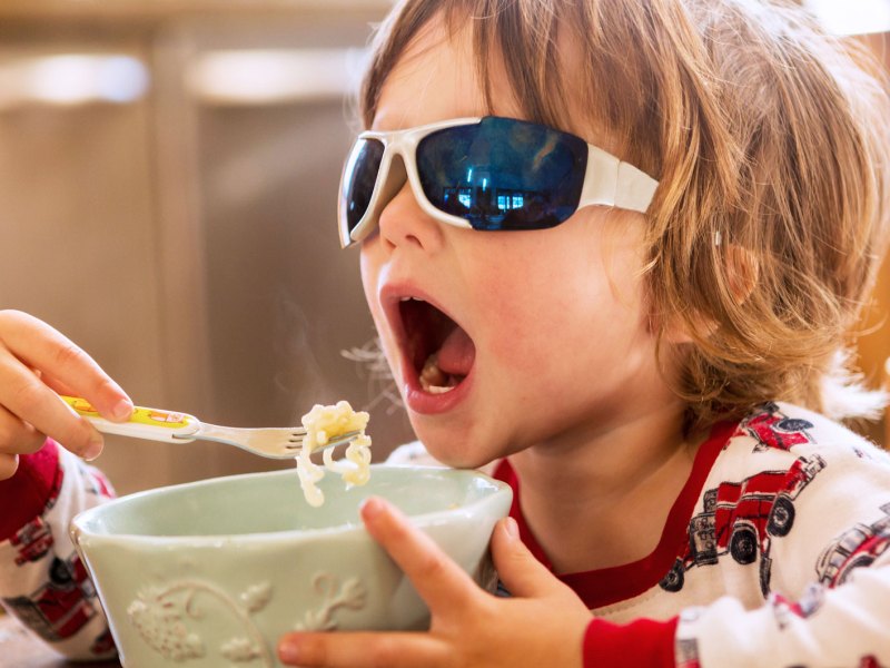 Kind, ca. 4 Jahre, sitzt mit Sonnenbrille am Frühstückstisch und isst sein Müsli.
