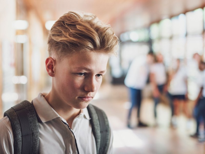 Männlicher Schüler, ca. 13 Jahre, geht mir gesenktem Kopf durch die Schule, im Hintergrund sieht man verschwommen andere Schüler, die scheinbar schlecht über ihn reden.